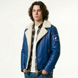 Könnte beinhalten: Blaue Lederjacke mit cremefarbenem Shearling-Kragen und -Futter. Die Jacke hat einen Aufnäher am Ärmel und wird über einem karierten Hemd und einem weißen T-Shirt getragen. Die Person trägt eine blaue Jeans.