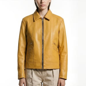 May include: Mustard yellow leather jacket with a black zipper closure and two front pockets. The jacket has a collared neckline and long sleeves. The model is wearing khaki pants.