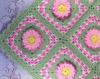 May include: A green and pink crocheted blanket with a floral pattern. The blanket is made with a repeating pattern of squares with pink flowers in the center.