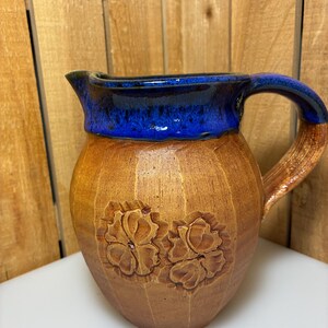 May include: A handcrafted ceramic pitcher with a brown body and a blue rim and handle. The pitcher features carved floral designs and vertical grooves. The handle has a brown and blue color scheme. This decorative pitcher is suitable for serving beverages.
