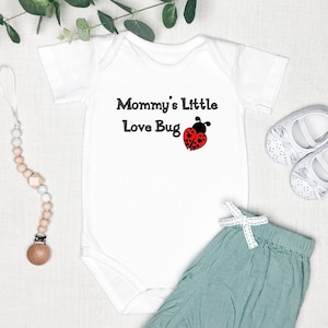 May include: White baby onesie with short sleeves and the text "Mommy's Little Love Bug" with a red ladybug graphic. The onesie is paired with a light green pair of trousers and a white wooden teether.