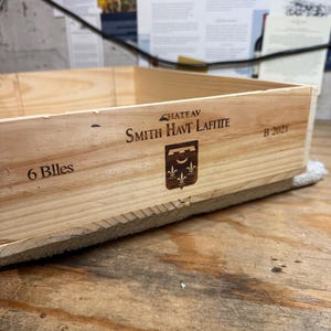 May include: A light-colored wooden wine crate featuring the black printed text CHATEAU SMITH HAVT LAFITTE, the year B 2021, the quantity 6 Blles, and a dark brown shield-shaped emblem with a crescent moon and three fleur-de-lis symbols.