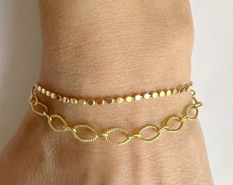 Può includere: Due braccialetti d'oro al polso di una persona. Il braccialetto superiore è una catena delicata con piccoli anelli rotondi. Il braccialetto inferiore è una catena più spessa con anelli ovali.