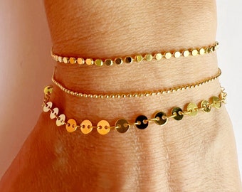 Può includere: Tre delicati braccialetti dorati sono mostrati su un polso. I braccialetti includono una catena di piccoli dischi rotondi, una catena di perline e una catena di dischi rotondi piatti più grandi. I braccialetti sono di colore oro lucido.