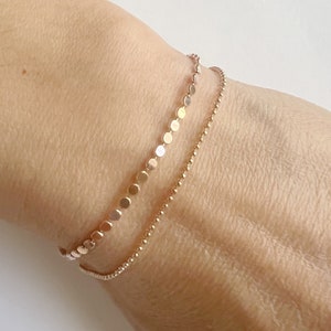 Puede incluir: Dos pulseras de oro en la muñeca de una persona. La pulsera superior está hecha de pequeños discos redondos. La pulsera inferior es una cadena delicada.