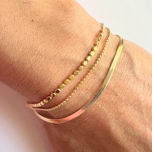 May include: Three gold bracelets stacked on a wrist. The top bracelet has small round beads, the middle bracelet has smaller round beads, and the bottom bracelet is a thin, flat, snake chain.