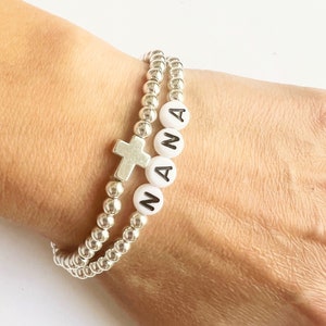 May include: A silver beaded bracelet with a silver cross charm and white beads spelling out the word "NANA".