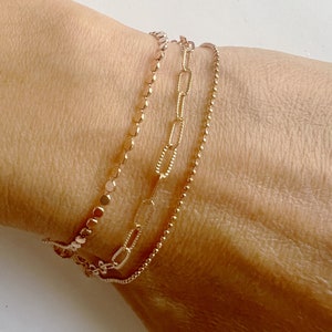 May include: Three delicate gold bracelets are stacked on a wrist. The bracelets feature different designs: a disc chain, a paperclip chain, and a beaded chain. The bracelets are a warm gold colour.