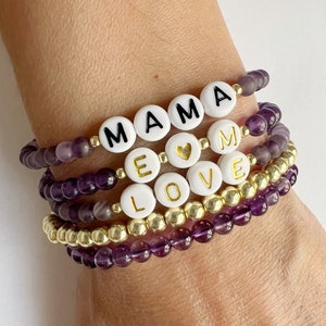 May include: Stack of beaded bracelets with the words "MAMA" and "LOVE" spelled out in white letter beads. The bracelets are purple and gold, with a gold heart charm.