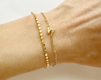 Può includere: Sono mostrati due braccialetti color oro. Uno è una catena di piccoli cerchi collegati. L'altro è una delicata catena con la lettera "W" come ciondolo. I braccialetti sono sul polso di una persona.