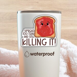 May include: A silver-colored tumbler featuring a sticker of a piece of toast with a smiling face covered in red jam. The sticker includes the text SPREAD TOO THIN AND STILL KILLING IT! The tumbler also displays a water droplet icon and the word waterproof.