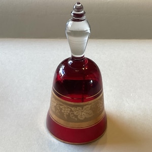 May include: A decorative ruby red glass bell with a clear handle. The bell features a gold band with a grape and leaf design. A vintage item, likely for display.