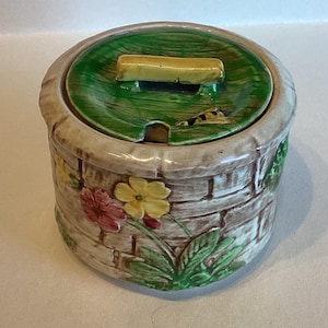 May include: A ceramic jar with a lid, featuring a brick-like pattern on the exterior. The lid is green with a yellow handle. Floral and leaf designs adorn the jar's sides. The jar is approximately 10 cm tall.