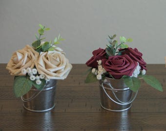 May include: Three small metal buckets with handles, each filled with artificial flowers. The buckets are silver and the flowers are ivory, burgundy, and a mix of ivory and brown.