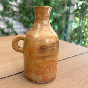 Puede incluir: Una botella de cerámica con cuello estrecho y una sola asa lateral, con un esmalte moteado en tonos naranja cálido, amarillo dorado y marrón tierra. La superficie tiene un acabado liso y brillante con sutiles variaciones de color.