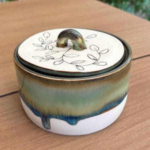 Puede incluir: Un tarro de cerámica redondo con tapa que presenta una base blanca con un efecto de esmalte de goteo verde y azul. La tapa es blanca con un patrón de enredadera de hojas negras dibujado a mano y un asa pequeña, curva y verdosa.
