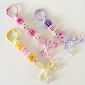 Può includere: Tre portachiavi personalizzati con perline a lettera in plastica colorata che compongono i nomi GRACE, ELLA e AMIRA. Ogni portachiavi include una chiusura in plastica, una perlina a forma di cuore o fiore, un fiocco in tessuto e perline.