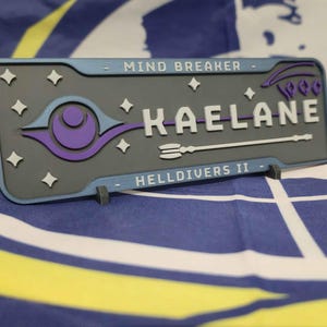 Op de afbeelding: Een rechthoekige naamplaat met de naam "KAELANE" in witte letters, met blauwe en paarse accenten. De tekst "MIND BREAKER" en "HELLDIVERS II" zijn ook zichtbaar. Geplaatst op een blauw en geel patroon.