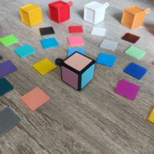 Può includere: Un piccolo cubo multicolore con una cornice nera. Il cubo ha un lato rosa chiaro, blu chiaro, viola chiaro e pesca chiaro. Il cubo è circondato da piccoli quadrati di vari colori, tra cui rosso, arancione, giallo, verde, blu, viola e grigio.