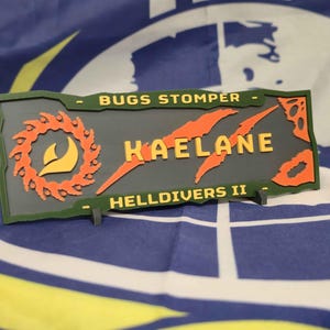 May include: A rectangular patch with the text "BUGS STOMPER", "KAELANE", and "HELLDIVERS II". The patch has a gray background with orange and green accents. The design includes a circular saw blade and claw marks.