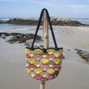 Può includere: Una borsa tote colorata in feltro con un motivo a forma di ventaglio nei toni del verde, giallo e viola. La borsa ha un bordo nero e manici neri. Lo sfondo mostra una spiaggia sabbiosa e l'oceano.