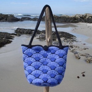 May include: A blue and black tote bag with a scalloped top and a pattern of overlapping blue and white scales. The bag is hanging from a wooden stick on a beach.