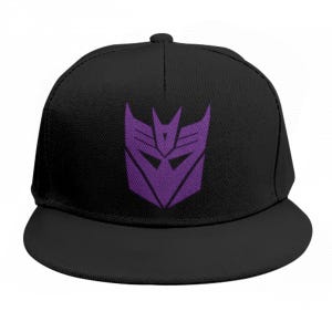 May include: A black baseball cap featuring a prominent purple Decepticon logo embroidered on the front panel. The hat has a structured crown, a flat brim, and a classic six-panel construction with a button at the top centre of the cap.