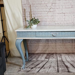 May include: A light blue console table with a white marble top and a single drawer. The table has four cabriole legs and a silver trim around the top edge. The table is sitting on a wood floor.