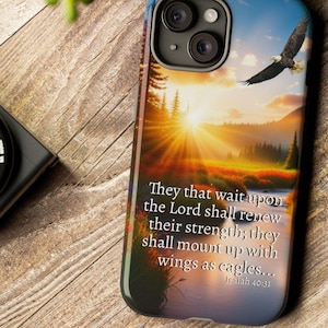 May include: A phone case featuring a scenic sunset with an eagle in flight. The case has a quote from Isaiah 40:31. The case is on a wooden surface with a camera and greenery in the background.