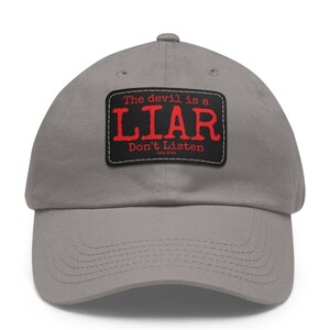 May include: A gray baseball cap with a curved brim. The cap features a black rectangular patch with the words "The devil is a LIAR Don't Listen" in red and white text. The text is a quote from John 8:44.