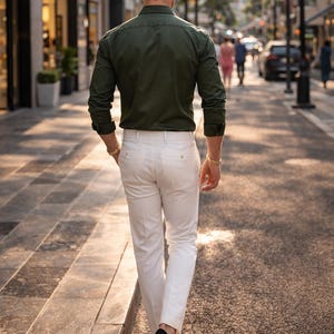 May include: A person wearing a dark green long-sleeve shirt and white pants walks down a city street. The person is wearing black shoes and gold bracelets. The background shows buildings, cars, and other people.