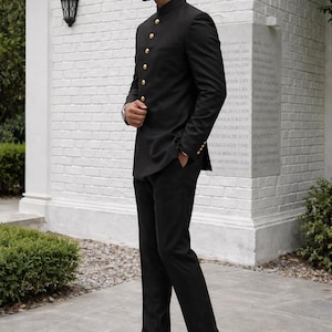 Puede incluir: Un traje negro con una chaqueta de cuello mandar&iacute;n y pantalones a juego. La chaqueta tiene botones dorados y mangas largas. El conjunto se combina con zapatos de vestir de cuero marr&oacute;n. El traje es un conjunto formal.