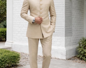 Puede incluir: Un traje beige con cuello mandarín, botones dorados y pantalón a juego. El traje se lleva con zapatos de cuero marrón oscuro y un reloj de pulsera plateado. La persona lleva gafas de sol negras.