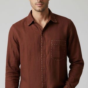 May include: A long-sleeved, brown button-down shirt with a chest pocket. The shirt features white stitching along the collar, pocket, and cuffs. The model is wearing the shirt with the top button undone.