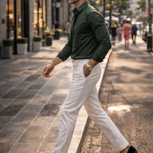 May include: A man in a dark green button-down shirt and white pants walks on a city street. He wears a gold watch and bracelet. Black loafers with gold embroidery complete the outfit. The background shows buildings and other pedestrians.