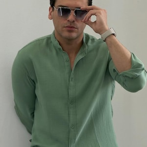 May include: A light green button-down shirt with a mandarin collar. The shirt is worn with sunglasses and a smartwatch. The shirt has long sleeves and is buttoned up. The person is leaning against a white wall.