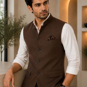 May include: A brown waistcoat with a mandarin collar and four dark buttons. The waistcoat has two front pockets and a pocket square. It is worn over a white dress shirt. The person is also wearing a black watch.