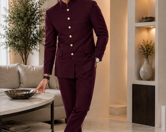 Puede incluir: Un traje burdeos con cuello mandarín y botones dorados. El conjunto incluye una chaqueta y un pantalón a juego. La chaqueta tiene un corte entallado y el pantalón es de corte ajustado. El atuendo se combina con zapatos de cuero marrón.