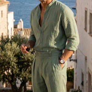 May include: A sage green long-sleeved shirt with a mandarin collar and matching sage green trousers. The shirt features a button-down front, and the trousers have a pleated waistline. A brown leather strap wristwatch is worn on the left wrist.