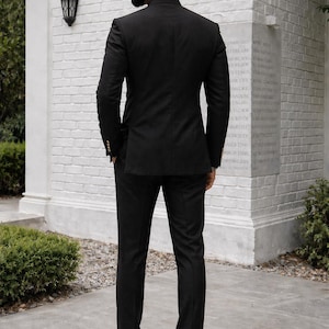 Puede incluir: Un traje negro con cuello mao, usado con zapatos de cuero marr&oacute;n. El traje tiene un corte entallado. El fondo incluye una pared de ladrillo blanco y un peque&ntilde;o arbusto verde. La chaqueta tiene botones dorados.