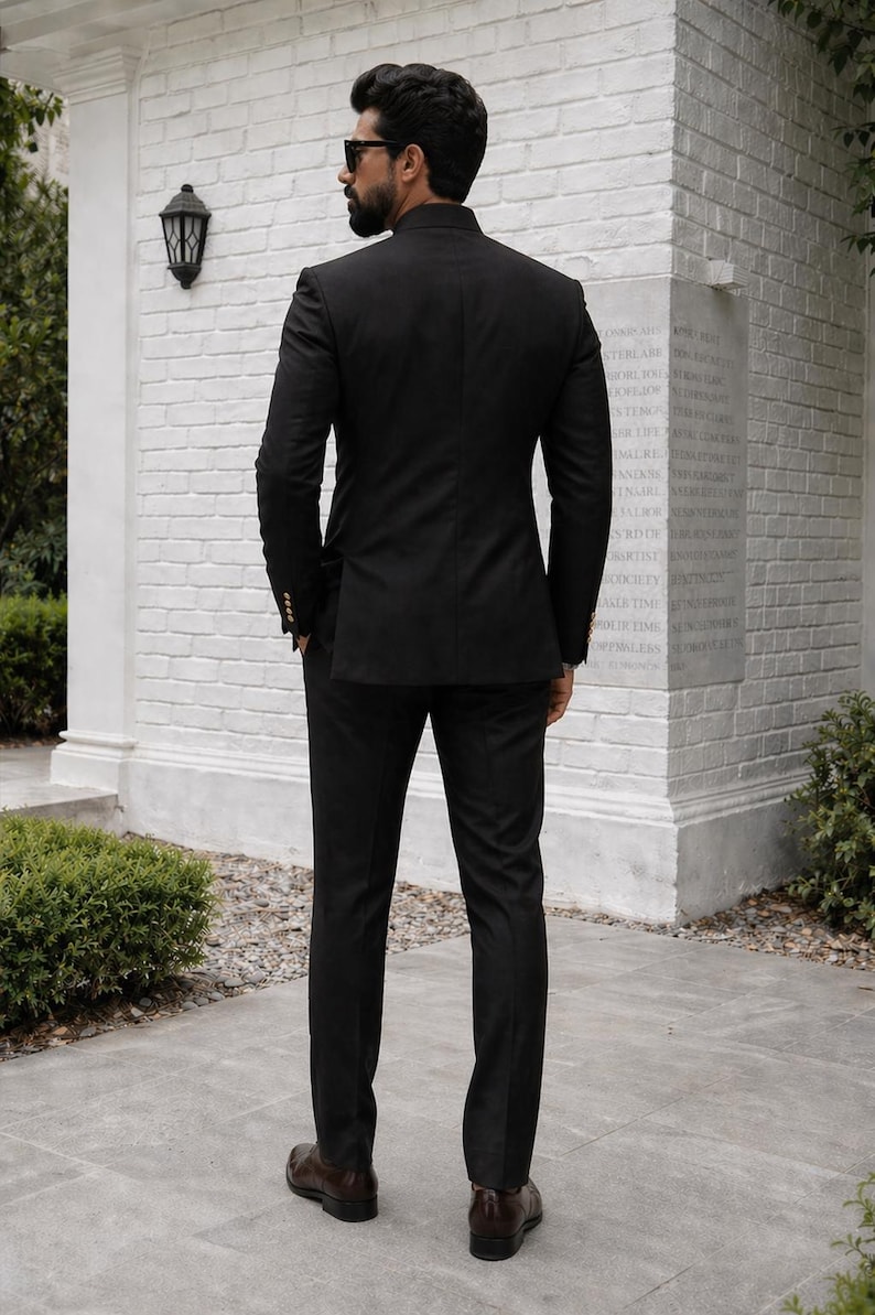 Puede incluir: Un traje negro con cuello mao, usado con zapatos de cuero marr&oacute;n. El traje tiene un corte entallado. El fondo incluye una pared de ladrillo blanco y un peque&ntilde;o arbusto verde. La chaqueta tiene botones dorados.