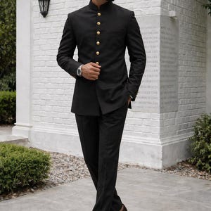 Puede incluir: Un traje negro con cuello mandar&iacute;n y botones dorados. El traje se combina con pantalones negros y zapatos de cuero marr&oacute;n. La persona lleva gafas de sol y un reloj. El fondo es una pared de ladrillos blancos.