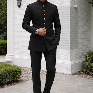 Puede incluir: Un traje negro con cuello mao, con botones dorados, combinado con pantalones negros y zapatos de cuero marr&oacute;n. El traje se lleva con gafas de sol y un reloj de pulsera. El escenario es al aire libre, con una pared de ladrillos blancos al fondo.