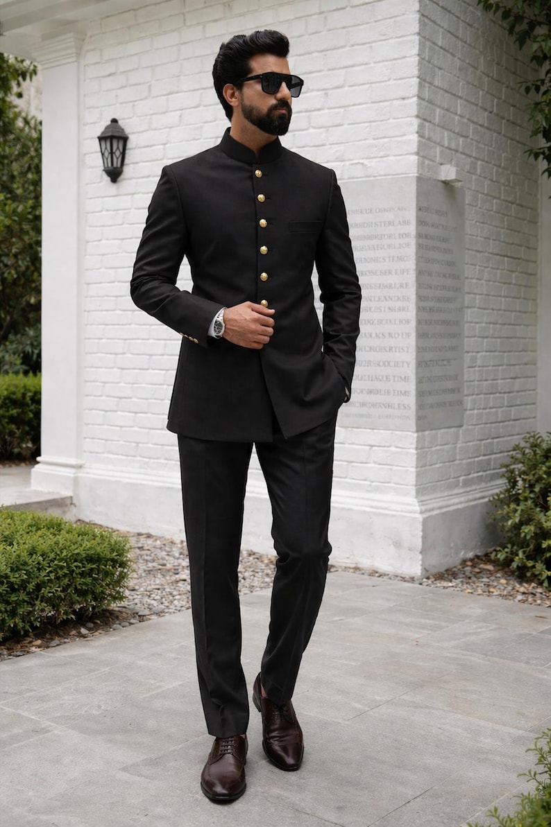 Puede incluir: Un traje negro con cuello mao, con botones dorados, combinado con pantalones negros y zapatos de cuero marr&oacute;n. El traje se lleva con gafas de sol y un reloj de pulsera. El escenario es al aire libre, con una pared de ladrillos blancos al fondo.