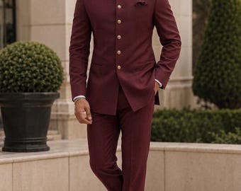 Puede incluir: Un traje bandhgala de color burdeos intenso que presenta una chaqueta estructurada con cuello mao, mangas largas y una hilera de botones dorados en la parte delantera. Los pantalones a juego tienen un corte recto y se combinan con zapatos negros.