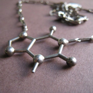 Puede incluir: Un collar de plata con un colgante con forma de mol&eacute;cula. El colgante est&aacute; hecho de peque&ntilde;as cuentas redondas conectadas por finas barras de metal.