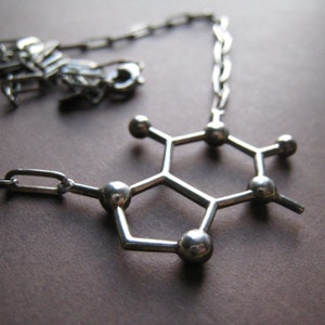 Puede incluir: Un collar de plata con un colgante en forma de mol&eacute;cule de cafe&iacute;na. El colgante est&aacute; hecho de metal y tiene un acabado brillante.