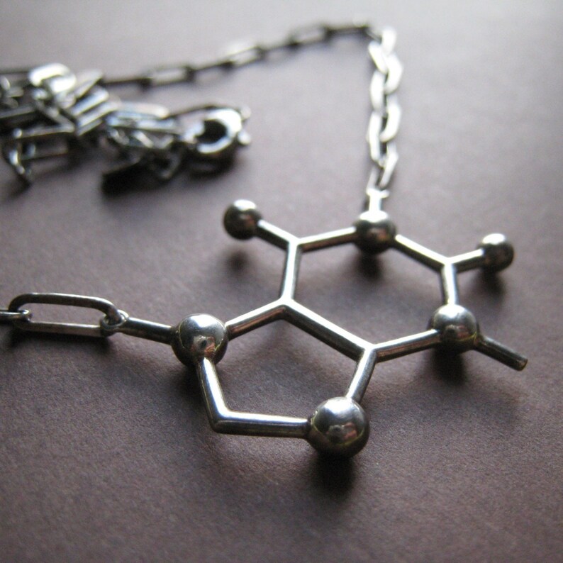 Puede incluir: Un collar de plata con un colgante en forma de mol&eacute;cule de cafe&iacute;na. El colgante est&aacute; hecho de metal y tiene un acabado brillante.