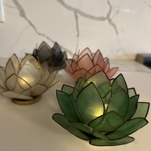 May include: Four lotus flower-shaped candle holders in various colors: white, gray, pink, and green. Each holder has a gold-colored trim and a small light inside, creating a warm glow. The candle holders are arranged on a white surface.