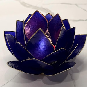 May include: A handcrafted, blue lotus flower candle holder with gold trim. The petals are a deep blue with a metallic sheen, and the interior is a lighter purple. The candle holder is designed to resemble a lotus flower.